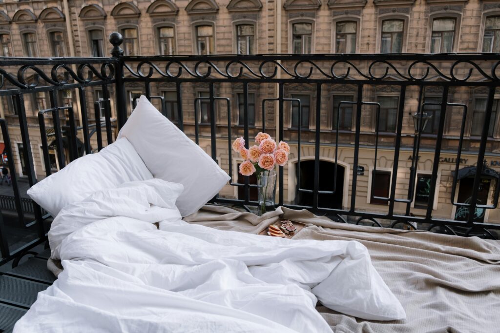Romantic balcony setting with white bed linens and pink roses overlooking an urban cityscape.